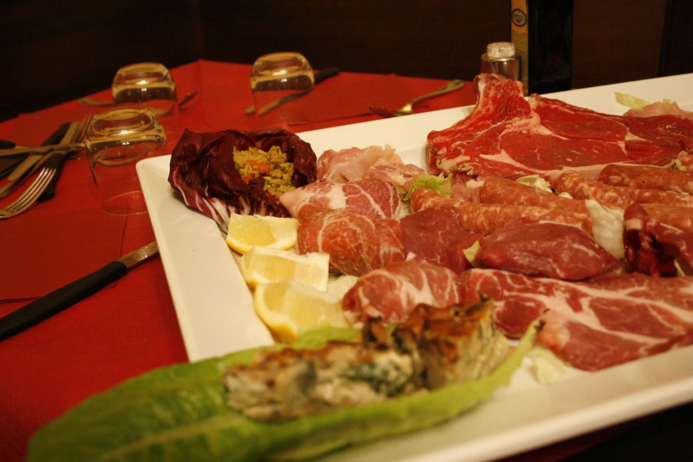Il vassoio della grigliata prima della cottura: braciole, capocollo, bocconcini di maiale, radicchio ripieno, spicchi di limone e un involtino di verdura su un piatto rettangolare bianco