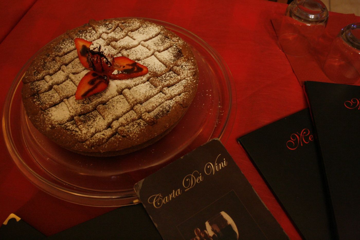 Crostata intera su alzata di vetro, con la griglia di pasta frolla spolverata di zucchero a velo e una fragola tagliata a ventaglio al centro