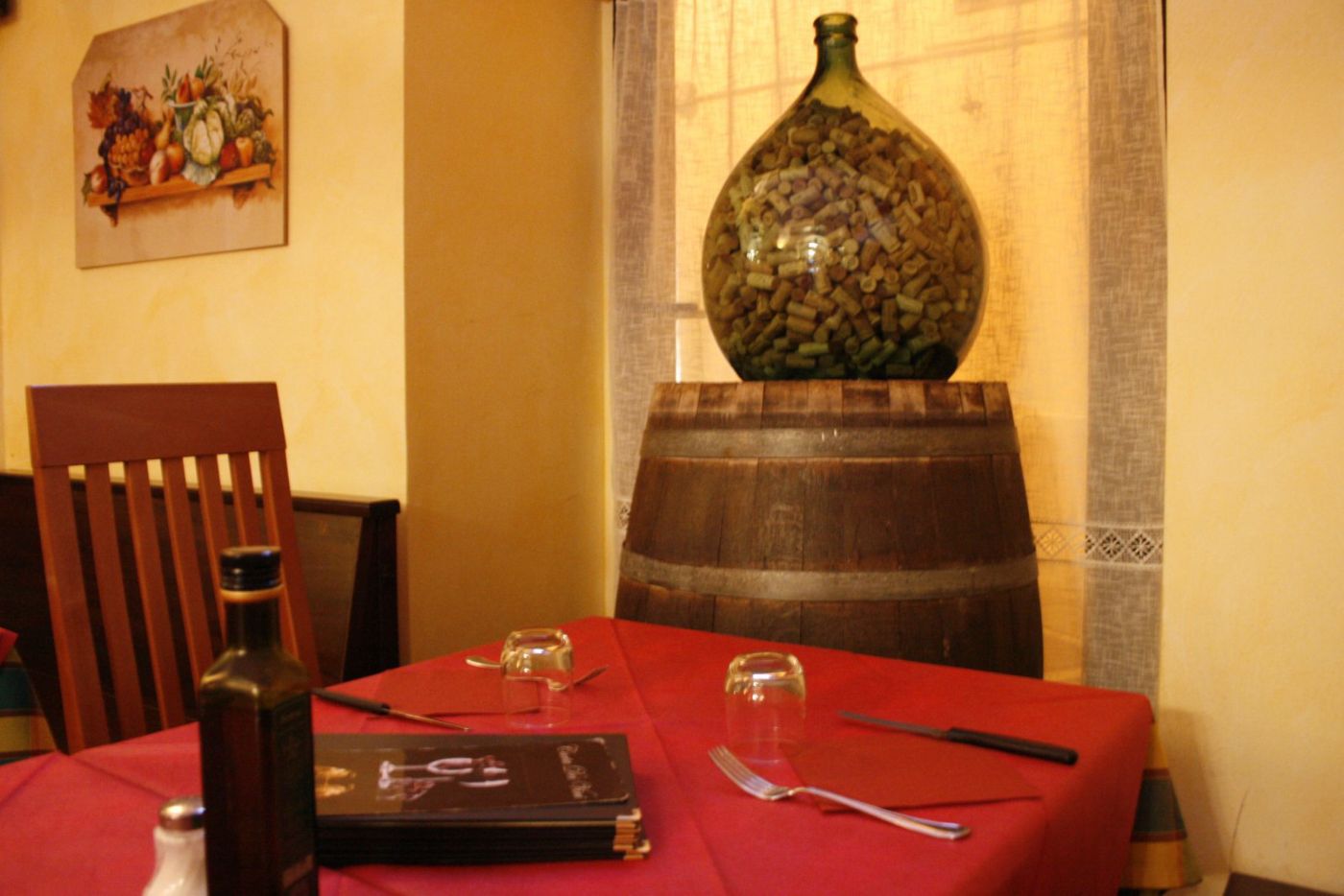 Damigiana di vetro verde piena di tappi di sughero appoggiata su una botte di legno, in un angolo della sala accanto a un tavolo apparecchiato