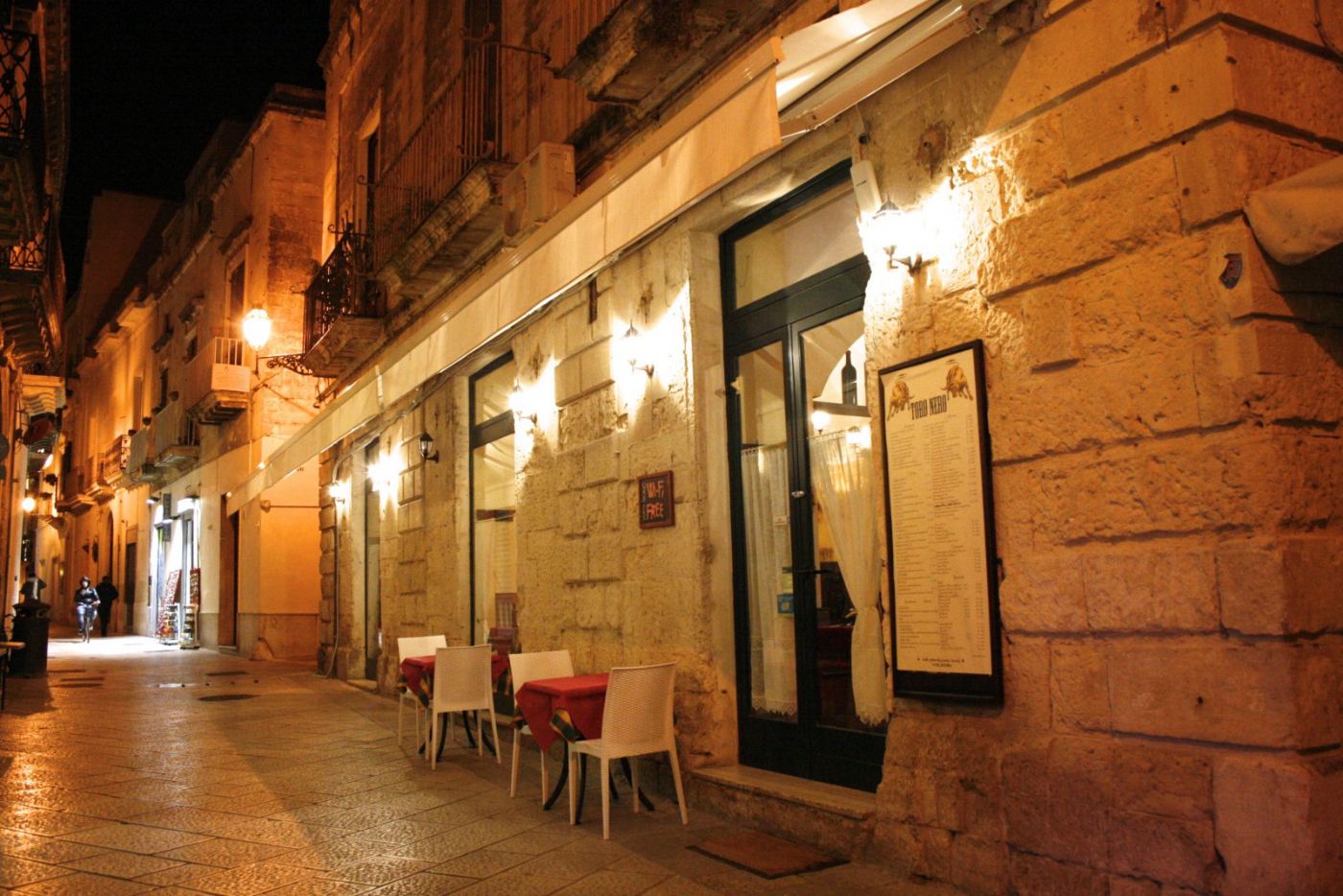 La facciata del Toro Nero di sera in via Federico d'Aragona: muro di pietra leccese illuminato dalle applique, tenda bianca, porta a vetri e il menu incorniciato accanto all'ingresso