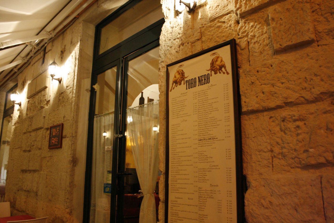 Il menu del Toro Nero incorniciato accanto alla porta d'ingresso, con il marchio dei due tori dorati in alto, sul muro di pietra leccese