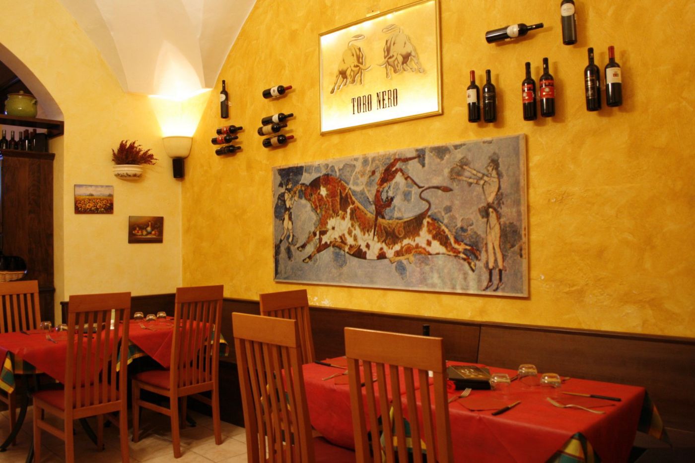 Sala interna del Toro Nero: parete ocra con l'insegna incorniciata in oro dei due tori, sotto una riproduzione dell'affresco minoico del salto del toro, bottiglie di vino appese a muro e tavoli con tovaglia rossa